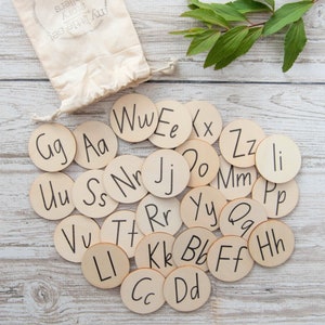 May include: A set of wooden alphabet letter tiles with a white background. The letters are written in black ink and include Aa, Bb, Cc, Dd, Ee, Ff, Gg, Hh, Ii, Jj, Kk, Ll, Mm, Nn, Oo, Pp, Qq, Rr, Ss, Tt, Uu, Vv, Ww, Xx, Yy, and Zz.
