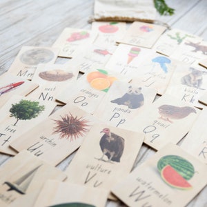 May include: Wooden alphabet flashcards with illustrations. Each card displays a letter, an image, and the word for the image. The images include a moon, tree, oranges, panda, and vulture. A small beige bag is in the background. The cards are made of wood.