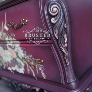 May include: Close-up of a dark purple dresser with floral accents and gold hardware. The dresser has ornate detailing, including carved leaf designs and decorative drawer pulls. The text "BRUSHED BY BRANDY" is visible.