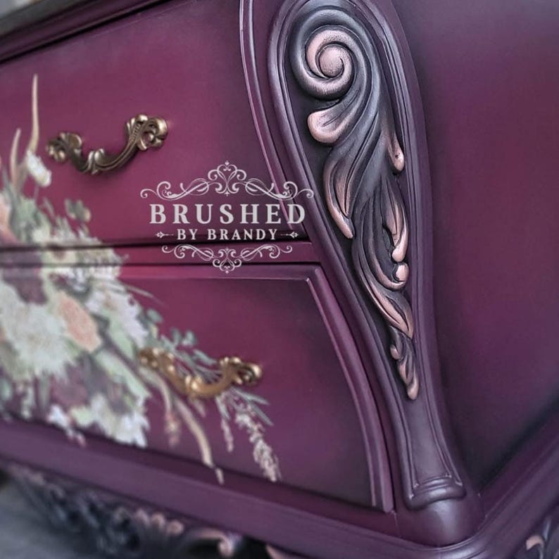May include: Close-up of a dark purple dresser with floral accents and gold hardware. The dresser has ornate detailing, including carved leaf designs and decorative drawer pulls. The text "BRUSHED BY BRANDY" is visible.