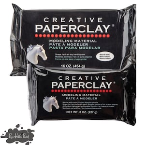 May include: Two black packages of Creative Paperclay modeling material. The packages display the brand name and product information in English, French, and Spanish. One package is labeled 16 oz. (454 g), and the other is 8 oz. (227 g). The packages feature a white unicorn.