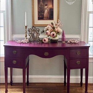 May include: A deep purple desk with four drawers and brass handles. The desk has a curved front and stands on four slender legs. A framed picture, flowers, and a candle holder adorn the top. The desk is in a room with a window.
