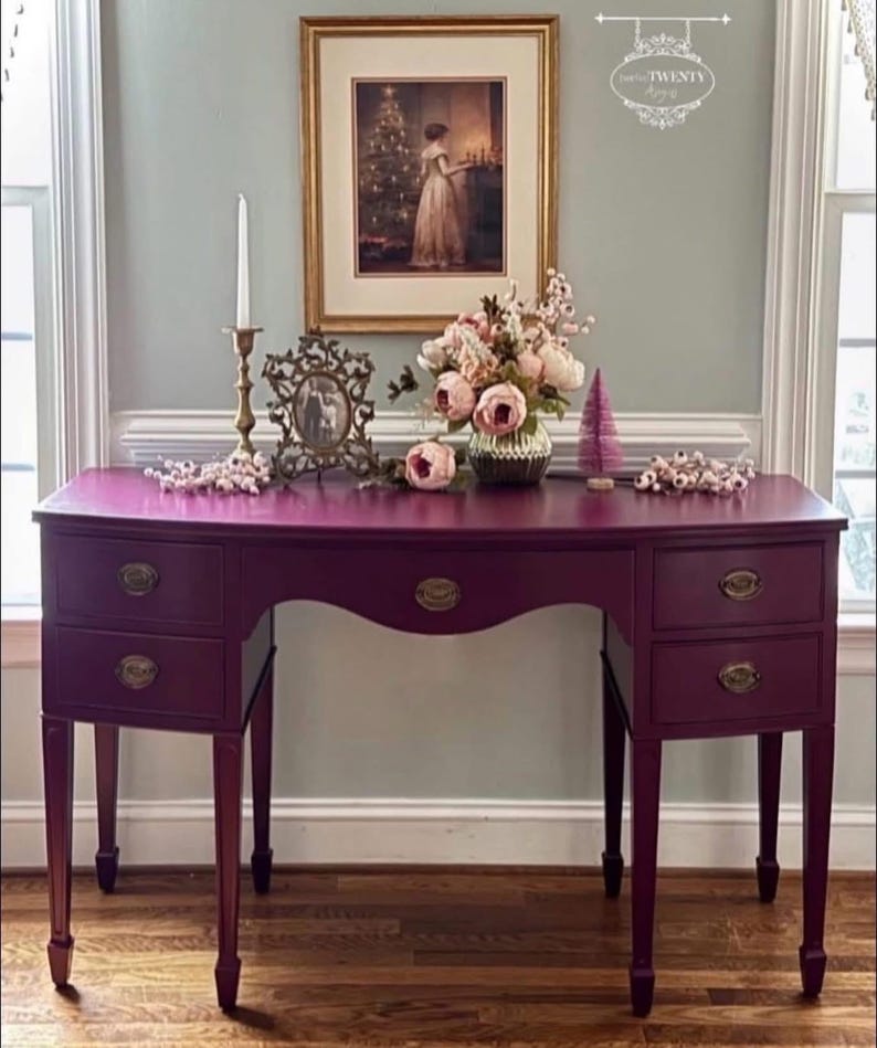 May include: A deep purple desk with four drawers and brass handles. The desk has a curved front and stands on four slender legs. A framed picture, flowers, and a candle holder adorn the top. The desk is in a room with a window.