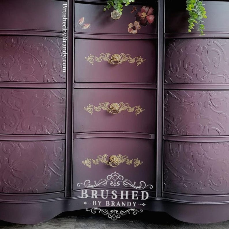 May include: A dark purple chest of drawers with gold handles and embossed floral patterns. The chest has multiple drawers and a curved design. The text "Brushed by Brandy" is visible at the bottom.