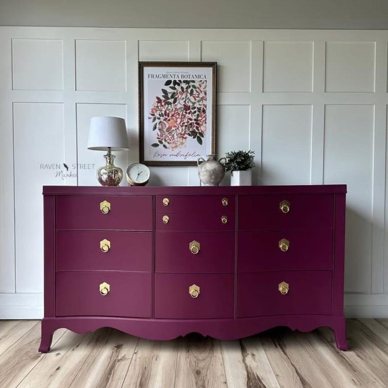 May include: A deep purple chest of drawers with six drawers and gold handles. It stands against a white panelled wall, with a botanical print in a frame and decorative items on top. The Raven Street Market logo is visible.
