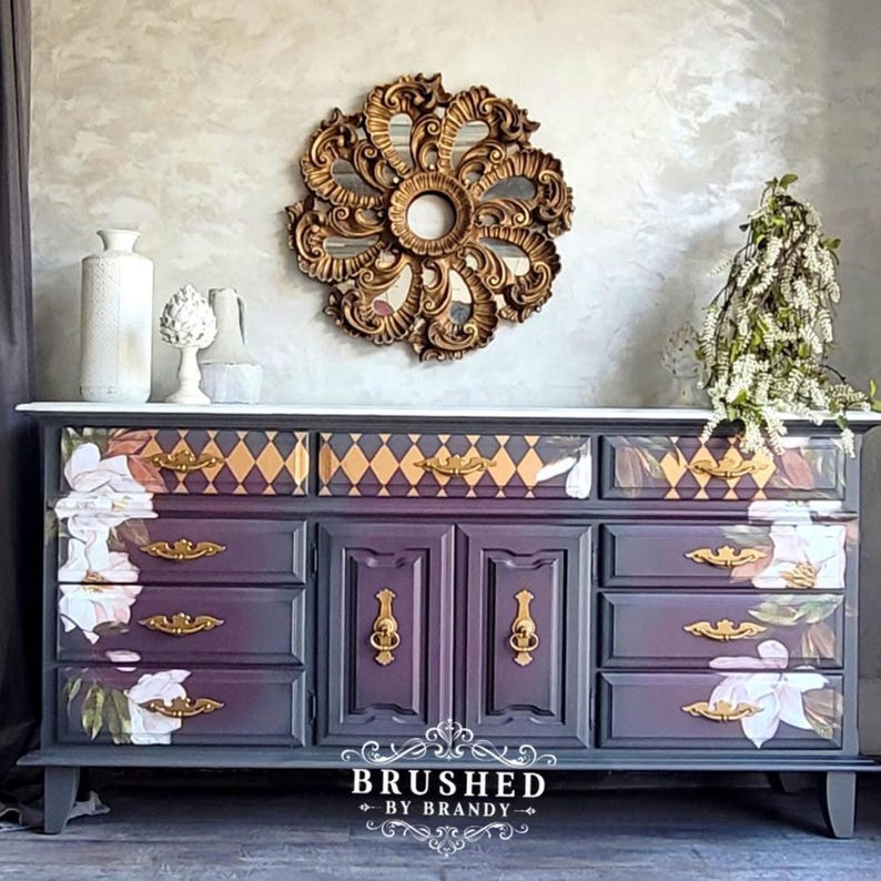May include: A hand-painted dresser in shades of purple and grey, with floral and geometric patterns. The dresser has multiple drawers and doors with gold hardware. A gold ornate mirror hangs above the dresser, and the text "BRUSHED by BRANDY" is visible.