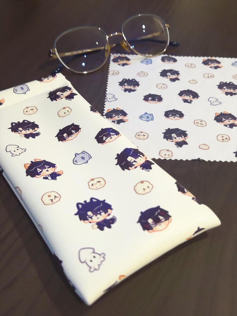 May include: Eyeglasses with gold frames rest on a white patterned cleaning cloth and a matching glasses case. The case and cloth feature a repeating pattern of cartoon characters and small, round, and ghost-like figures.