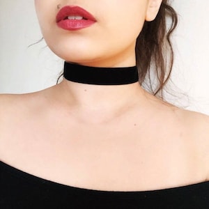 May include: A black velvet choker necklace. The choker is a simple band of black velvet, worn close to the neck. The image shows the choker against a person's skin, with a black top and a neutral background.
