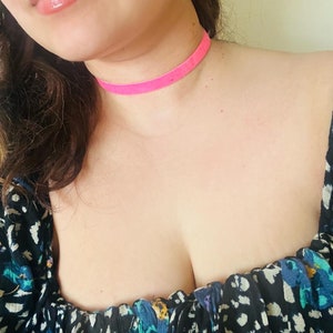 May include: A close-up shot of a person wearing a bright pink velvet choker necklace. The person is also wearing gold hoop earrings and a black top with a blue and white floral pattern.