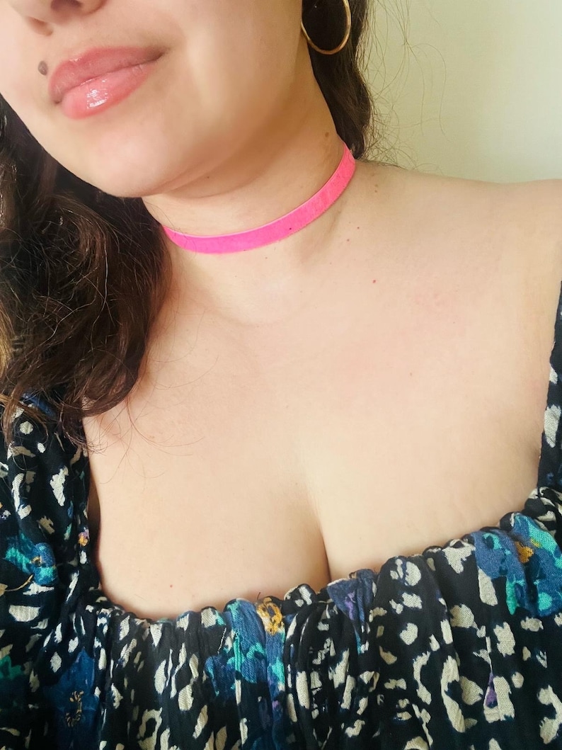 May include: A close-up shot of a person wearing a bright pink velvet choker necklace. The person is also wearing gold hoop earrings and a black top with a blue and white floral pattern.