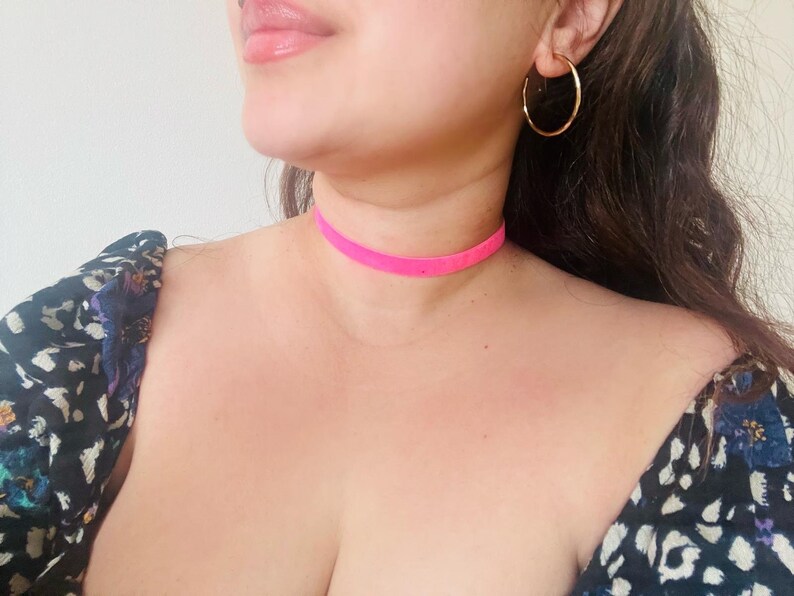 May include: A close-up shot of a person wearing a bright pink choker necklace. The necklace is a thin band of fabric. The person is also wearing gold hoop earrings and a floral print top.