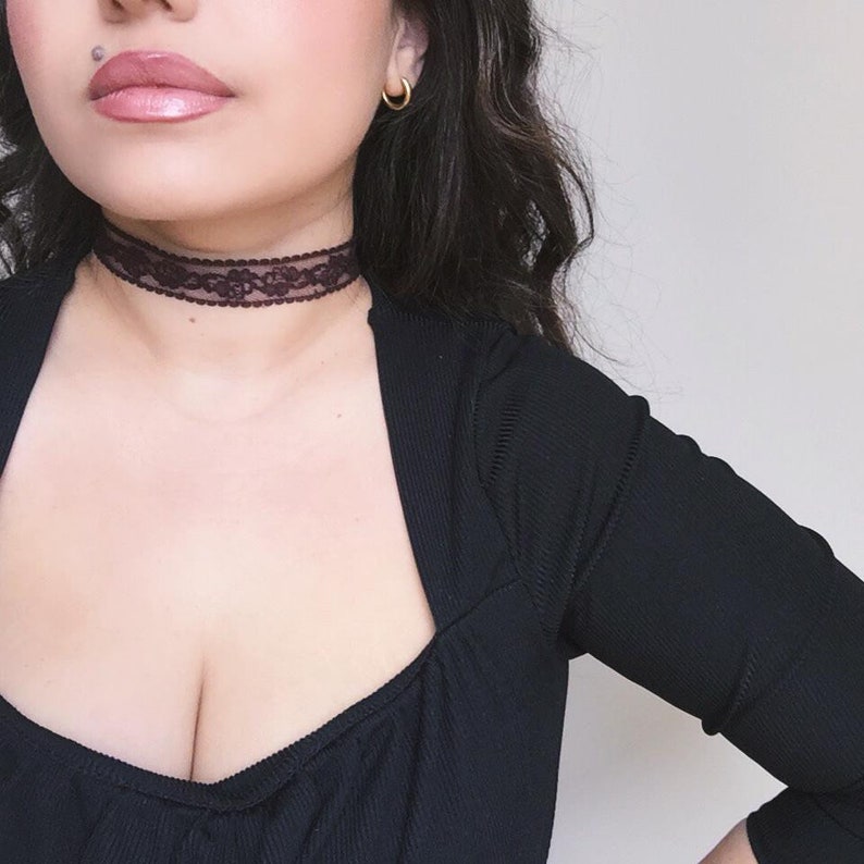 May include: A dark burgundy lace choker necklace. The choker is worn around the neck. The model is wearing a black top and gold hoop earrings. The background is a plain, light colour.