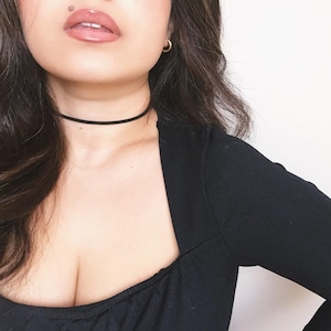 May include: A black choker necklace is worn around the neck. The person is wearing a black square-neck top and gold hoop earrings. The person has dark hair and is wearing lip gloss.