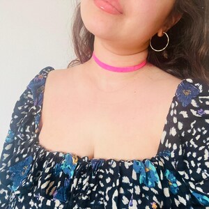 May include: A bright pink choker necklace is worn around the neck. The person is wearing a black top with a white and blue floral pattern and gold hoop earrings. The background is a plain white wall.