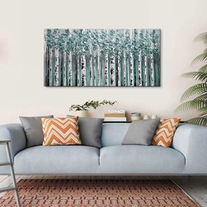 Op de afbeelding: Een rechthoekig canvas schilderij toont een bos van berken met witte stammen en turquoise bladeren. Het kunstwerk hangt boven een lichtblauwe bank, versierd met grijze en oranje kussens met patronen. Een vloerkleed met patroon en een potplant zijn ook zichtbaar.