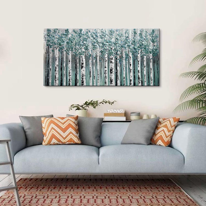 Op de afbeelding: Een rechthoekig canvas schilderij toont een bos van berken met witte stammen en turquoise bladeren. Het kunstwerk hangt boven een lichtblauwe bank, versierd met grijze en oranje kussens met patronen. Een vloerkleed met patroon en een potplant zijn ook zichtbaar.
