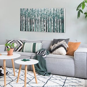 Op de afbeelding: Een abstract canvas kunstwerk dat een bos van berkenbomen met getextureerde turquoise en witte bladeren afbeeldt. Het kunstwerk hangt boven een grijze bank met decoratieve kussens en een groene deken. Twee witte bijzettafels met houten poten staan voor de bank.