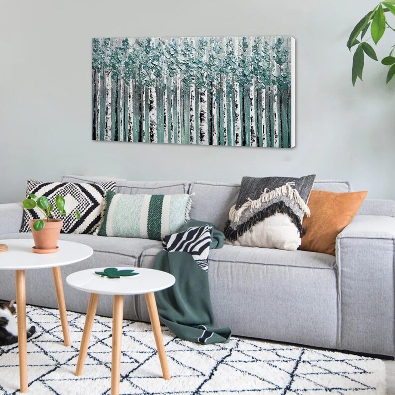 Op de afbeelding: Een abstract canvas kunstwerk dat een bos van berkenbomen met getextureerde turquoise en witte bladeren afbeeldt. Het kunstwerk hangt boven een grijze bank met decoratieve kussens en een groene deken. Twee witte bijzettafels met houten poten staan voor de bank.