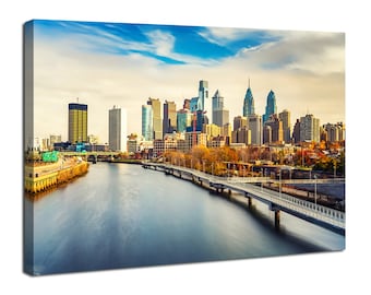 May include: A panoramic view of the Philadelphia skyline with a river in the foreground and a bridge leading to the city. The buildings are tall and modern, with a mix of glass and brick. The sky is blue with white clouds.