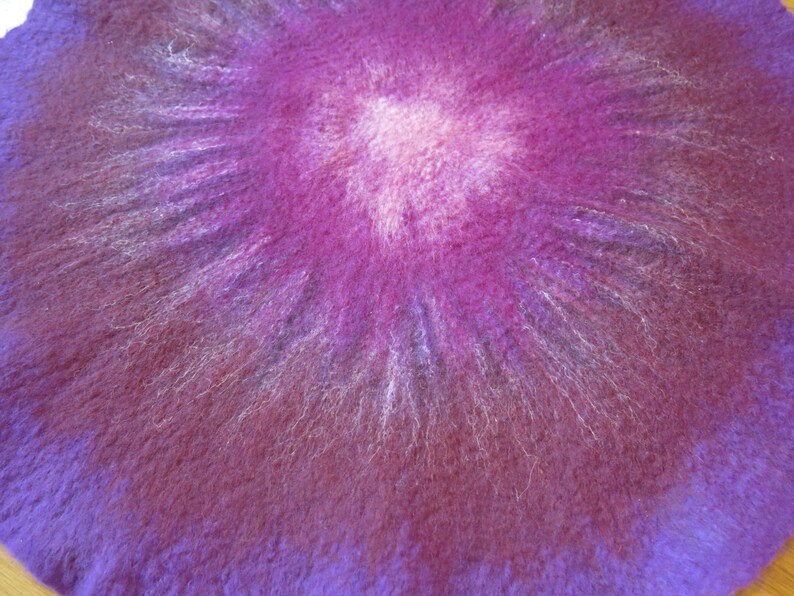 Puede incluir: Una pieza textil circular de fieltro con aguja que presenta un degradado de colores vibrantes. El centro es de un suave rosa p&aacute;lido que transiciona hacia afuera en tonos magenta, morado intenso y violeta, con una superficie fibrosa y texturizada.