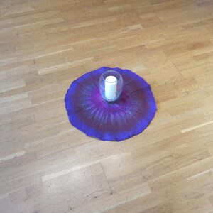 Puede incluir: Una alfombrilla circular de lana fieltrada con un degradado vibrante de tonos p&uacute;rpura intenso y magenta, que irradia hacia afuera desde el centro. Un portavelas de cristal transparente con una vela de pilar blanca se sit&uacute;a en el centro.