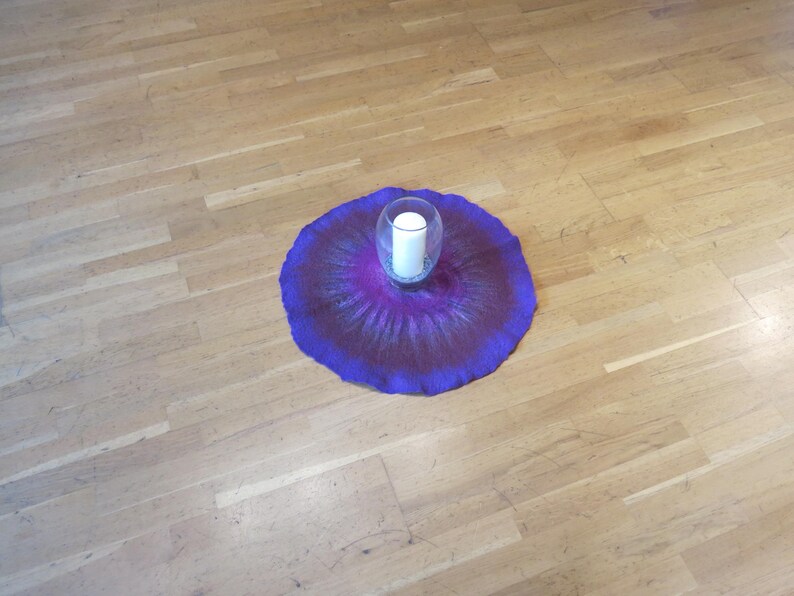 Puede incluir: Una alfombrilla circular de lana fieltrada con un degradado vibrante de tonos p&uacute;rpura intenso y magenta, que irradia hacia afuera desde el centro. Un portavelas de cristal transparente con una vela de pilar blanca se sit&uacute;a en el centro.