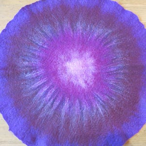 Puede incluir: Una alfombrilla circular de lana fieltrada con un degradado de colores vibrantes. El centro es de un rosa claro suave que transiciona a magenta profundo, luego a p&uacute;rpura intenso y finalmente a un borde violeta oscuro y fibroso.