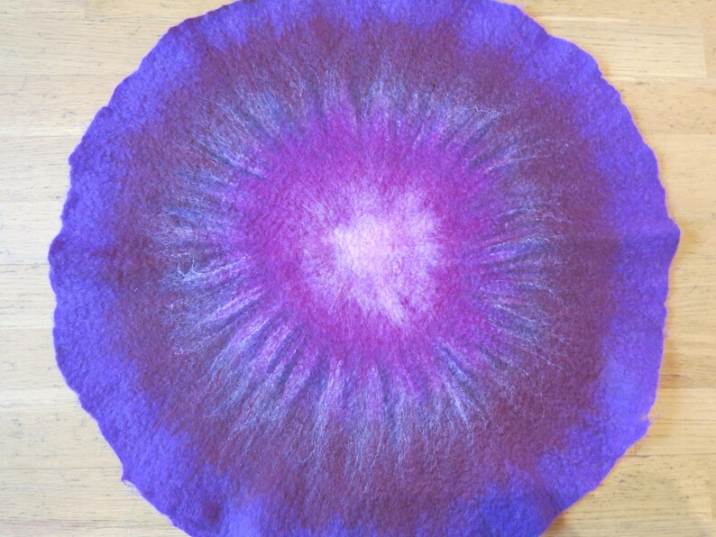Puede incluir: Una alfombrilla circular de lana fieltrada con un degradado de colores vibrantes. El centro es de un rosa claro suave que transiciona a magenta profundo, luego a p&uacute;rpura intenso y finalmente a un borde violeta oscuro y fibroso.