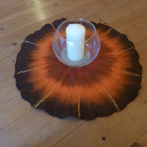 May include: A round, brown and orange felted wool rug with gold accents. A clear glass bowl with a white candle sits in the center of the rug.