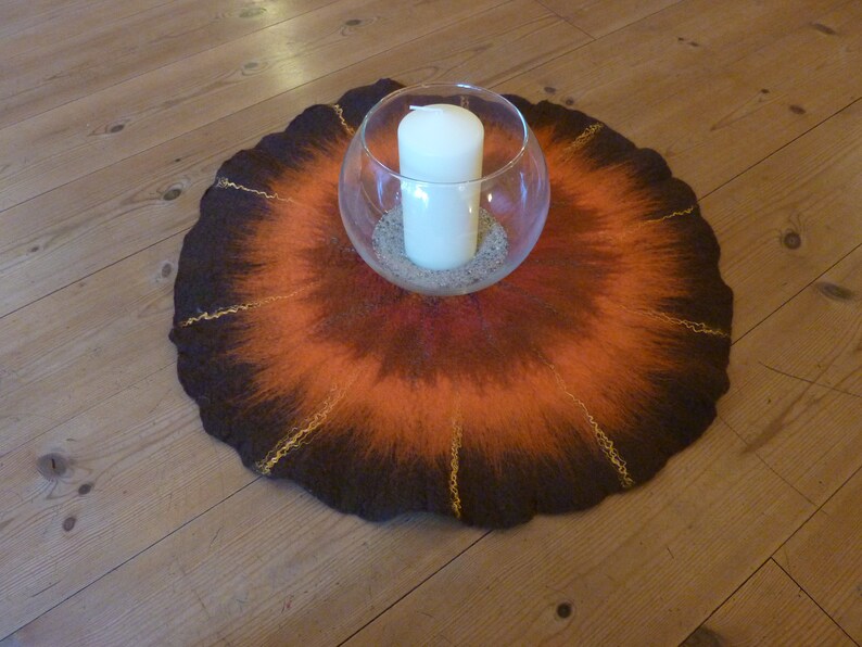 May include: A round, brown and orange felted wool rug with gold accents. A clear glass bowl with a white candle sits in the center of the rug.