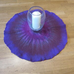 Puede incluir: Un tapete de fieltro circular con un degradado vibrante en tonos morado, magenta y azul oscuro. Centrado en el tapete hay un jarr&oacute;n de vidrio transparente que contiene una vela de pilar blanca sobre una base de peque&ntilde;as piedras decorativas grises.