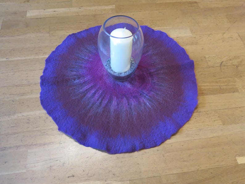 Puede incluir: Un tapete de fieltro circular con un degradado vibrante en tonos morado, magenta y azul oscuro. Centrado en el tapete hay un jarr&oacute;n de vidrio transparente que contiene una vela de pilar blanca sobre una base de peque&ntilde;as piedras decorativas grises.
