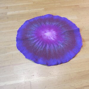 Puede incluir: Una alfombrilla circular de fieltro con un degradado de colores vibrantes que transita desde un centro blanco brillante a tonos magenta, morado intenso y un borde exterior violeta oscuro, con superficie texturizada y borde ondulado.