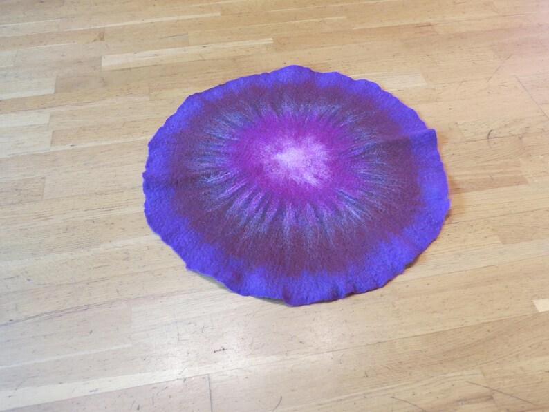 Puede incluir: Una alfombrilla circular de fieltro con un degradado de colores vibrantes que transita desde un centro blanco brillante a tonos magenta, morado intenso y un borde exterior violeta oscuro, con superficie texturizada y borde ondulado.
