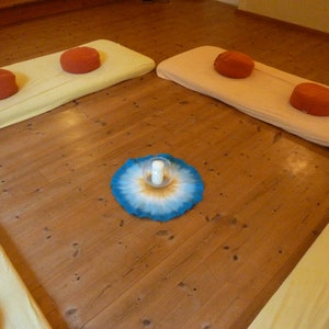 May include: A wooden floor with four cushions, two yellow and two orange, arranged in a square. A blue and white mandala with a glass candle holder in the center is on the floor.