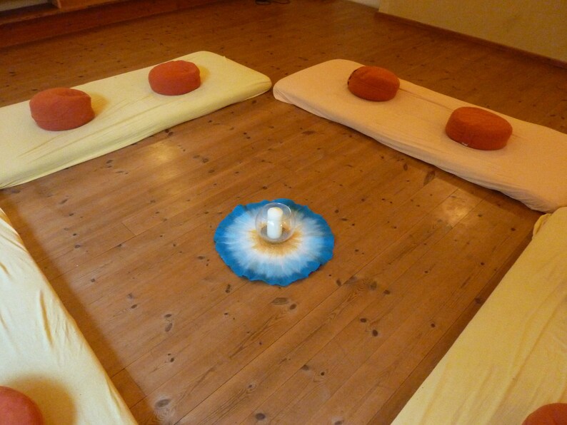 May include: A wooden floor with four cushions, two yellow and two orange, arranged in a square. A blue and white mandala with a glass candle holder in the center is on the floor.