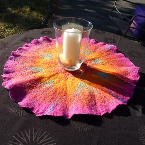 Puede incluir: Un tapete redondo de lana afieltrada con un degradado vibrante de rosa intenso y naranja brillante, acentuado con detalles de fibras azules radiantes. Un portavelas de cristal transparente con una vela blanca se sit&uacute;a en el centro.
