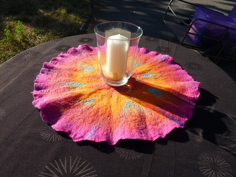 Puede incluir: Un tapete redondo de lana afieltrada con un degradado vibrante de rosa intenso y naranja brillante, acentuado con detalles de fibras azules radiantes. Un portavelas de cristal transparente con una vela blanca se sit&uacute;a en el centro.