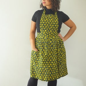 May include: A woman wearing a black turtleneck top and a yellow and black patterned apron with a pocket. The apron has a gathered skirt.