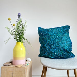 May include: A yellow and pink ceramic vase with a bouquet of purple flowers sits on a wooden table. A blue and green patterned throw pillow sits on a gray chair.