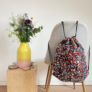 Könnte beinhalten: Eine rote und blaue Blumendrawstring-Tasche mit schwarzen Kordeln. Die Tasche sitzt auf einem grauen Stuhl mit einer gelben und pinkfarbenen Vase mit Blumen im Hintergrund.