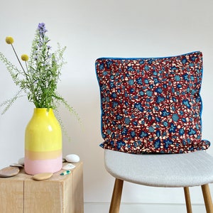 May include: A yellow and pink ceramic vase with a bouquet of purple and yellow flowers sits on a wooden table next to a gray chair with a red, blue, and white patterned cushion.