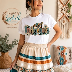 Könnte beinhalten: Ein weißes T-Shirt mit dem Wort "Indigenous" in bunten Buchstaben und einem Schildkröten-Motiv. Das Outfit umfasst einen cremefarbenen Rock mit türkisfarbenen, orangefarbenen und weißen Streifen und einem türkisfarbenen Saum. Das Bild enthält auch den Text "crafted by Lashell".