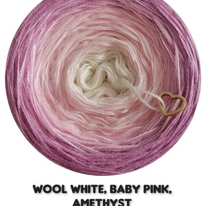 Op de afbeelding: Een ronde bol garen in een kleurengradatie, waaronder wit, babyroze en amethist. Een houten hartvormige bedel is aan het garen bevestigd. De tekst "WOOL WHITE, BABY PINK, AMETHYST" staat onderaan.