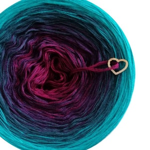 May include: A vibrant ball of yarn in teal, purple, and magenta, with a wooden heart-shaped charm. The yarn's colours blend from teal to purple to magenta. Suitable for knitting and crochet.
