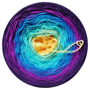May include: A skein of yarn in a gradient of purple, blue, turquoise, and yellow colours. The yarn is wound into a circle and the centre of the circle is visible. A wooden heart tag is attached to the yarn.