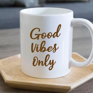 May include: White ceramic mug with the words "Good Vibes Only" in a brown script. The mug sits on a light brown wooden tray. The mug has a curved handle.