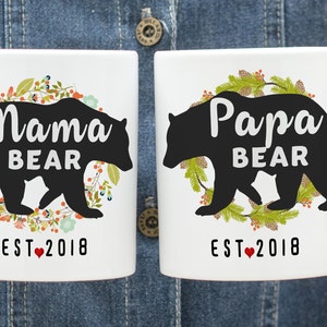 May include: Two white ceramic mugs with black bear silhouettes and floral wreaths. The mugs read "Mama Bear Est. 2018" and "Papa Bear Est. 2018".