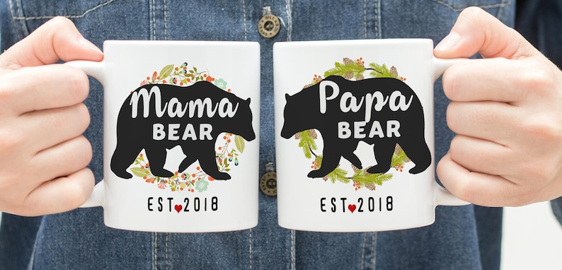 May include: Two white ceramic mugs with black bear silhouettes and floral wreaths. The mugs read "Mama Bear Est. 2018" and "Papa Bear Est. 2018".