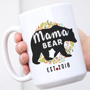 May include: White ceramic mug featuring a black silhouette of a bear and the text "Mama Bear Est. 2018" encircled by a floral wreath.